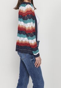 Sweter kardigan z falistym, wielokolorowym wzorem w kolorach czerwonym, turkusowym, białym i granatowym. Posiada otwarty przód oraz teksturowany, lekki materiał.