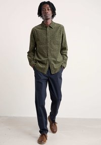 Camicia a maniche lunghe verde a fantasia con colletto, abbinata a pantaloni scuri e scarpe marroni. La camicia presenta un motivo geometrico.