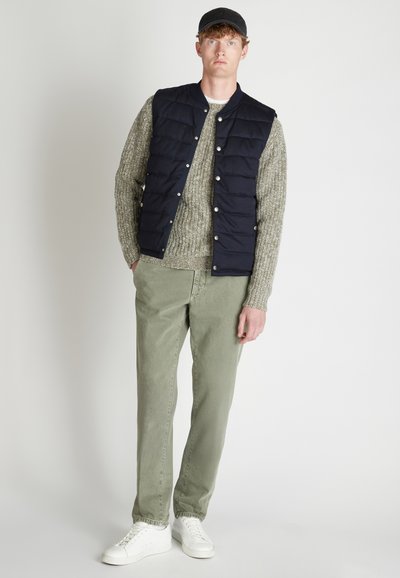 Marineblauwe puffer vest met drukknoopjes over een textuurbeige trui, gecombineerd met lichtgroene broek en witte sneakers. Een simpele pet maakt de look compleet.