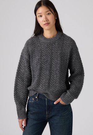 Femme aux longs cheveux foncés portant un pull en maille texturé gris foncé et un jean bleu, debout avec une main dans la poche devant un fond uni.
