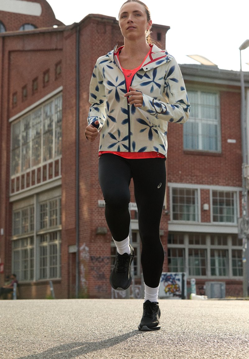 Sportjacke mit Blumenmuster in Creme und Marineblau, kombiniert mit einem roten Oberteil, schwarzen Leggings und schwarzen Laufschuhen. Außenbereich.