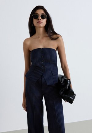 Donna che indossa un top senza spalline a righe blu navy e pantaloni abbinati, tiene una borsa in pelle nera e indossa occhiali da sole scuri su uno sfondo uniforme.