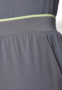 Grå atletisk klädesplagg med en slät textur, med en bred midjeband som framhävs av en tunn grön accent. Lätt rynkdetalj i botten.