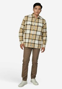 Junger Mann stehend, trägt ein braun-weiß kariertes Hemd mit Knöpfen, braune Hose und weiße Turnschuhe vor einem einfarbigen hellen Hintergrund.