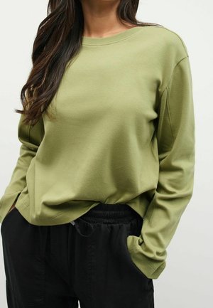 Femme portant une chemise à manches longues vert olive légèrement rentrée dans un pantalon noir à taille élastique, les mains dans les poches.