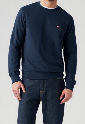 Man draagt een marineblauwe Levi's sweatshirt met lange mouwen en donkere spijkerbroek, staand tegen een effen lichte achtergrond.