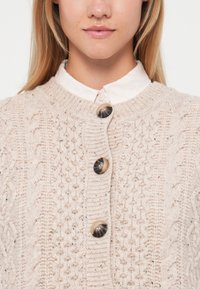 Světle růžový pletený kardigan s krajkovými vzory, vybavený velkými tmavými knoflíky. Hladový límec a textura ukazují na měkkou, silnou přízi.