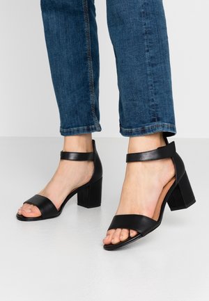 Fødder iført sorte læder åbne tå blokhæl sandaler med ankelremme, kombineret med blå cropped denim jeans på en hvid gulv.