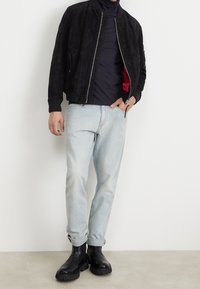 Giacca bomber nera in suede con polsini a coste, tasche con zip e un rivestimento rosso, abbinata a jeans blu chiaro e stivaletti neri.