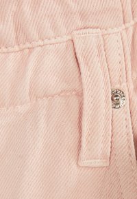 Ljusrosa denimtyg med en synlig ficka, kontrasterande sömmar och en silverfärgad tryckknapp. Visar textur och vävdetaljer.