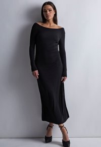 Robe longue noire à épaules dénudées fabriquée dans un tissu lisse et extensible. Présente des manches longues et une fente haute sur un côté. Associée à des chaussures à talons noires.