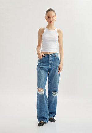 Top sin mangas de canalé blanco y pantalones vaqueros anchos de mezclilla azul con rotos. La tela parece suave y la mezclilla está desgastada en las rodillas. Bailarinas negras con tachuelas plateadas.