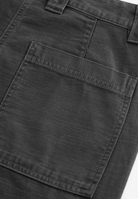 Gros plan sur une poche de pantalon en tissu noir avec des coutures visibles et une passants de ceinture au-dessus de la poche.