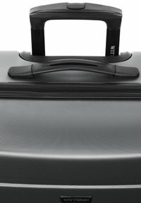 Black hardshell suitcase with textured surface, retractable black handle, and zip closure. Features a logo plate on the front.
