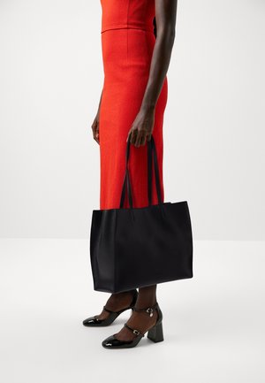 Kobieta w czerwonej sukience trzymająca dużą czarną skórzaną torbę typu tote, nosząca czarne lakierowane buty na niskim obcasie typu Mary Jane, stojąca na białej podłodze.