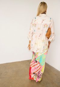 Γυναίκα με μακριά ξανθά μαλλιά, που φοράει ανοιχτόχρωμο πουκάμισο με πολύχρωμα δυτικά μοτίβα και ζωντανή πολύχρωμη μάξι φούστα, κρατώντας ένα καφέ πορτοφόλι.