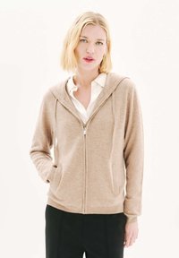 Sudadera beige con cremallera fabricada en un tejido suave, que cuenta con una cremallera frontal, dos bolsillos laterales y una capucha. Se lleva sobre una camisa blanca con cuello.