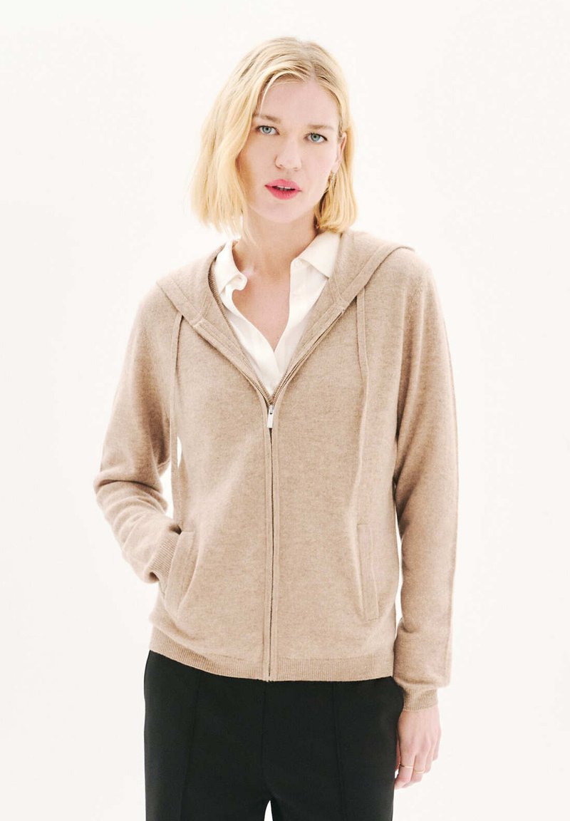 Sudadera beige con cremallera fabricada en un tejido suave, que cuenta con una cremallera frontal, dos bolsillos laterales y una capucha. Se lleva sobre una camisa blanca con cuello.