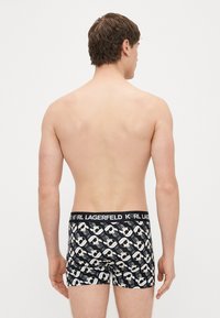 Melnas boksa šorti ar Karl Lagerfeld logotipa lenti, uz kuras atkārtoti attēlotas stilizētas galvas krēmkrāsā un pelēkā krāsā.