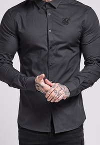 Camisa negra de botones con mangas largas, hecha de tela suave, que presenta un pequeño logotipo bordado en el pecho y puños abotonados.