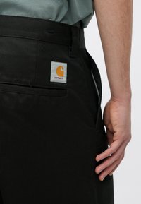 Schwarze Hosen aus strapazierfähigem Material verfügen über eine Gesäßtasche und ein sichtbares Carhartt-Logo-Patch in Orange auf weißem Hintergrund.