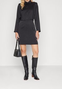 Svart klänning med knäppkrage, långa ärmar och en figursydd silhuett. Matchad med knähöga svarta läderstövlar och en liten handväska.