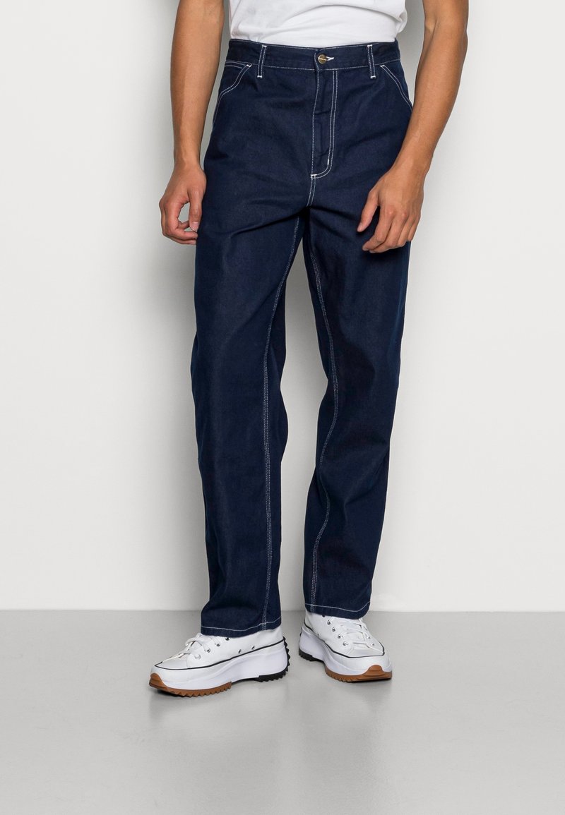 Pantalones vaqueros de mezclilla azul oscuro con cintura alta, costuras blancas y bolsillos delanteros. Combinados con zapatillas blancas que tienen suela marrón y negra.