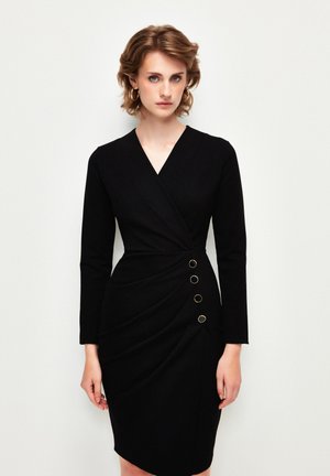 Abito avvolgente nero con maniche lunghe, caratterizzato da una scollatura a V, una texture drappeggiata e cinque dettagli con anelli dorati sul lato. Finitura in tessuto liscia.