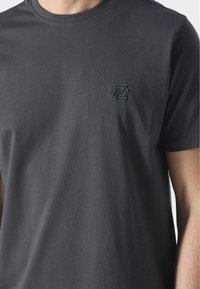 Férfi rövid ujjú póló sötét szürke pamutból. Kerek nyakkivágással és dombornyomott logóval a bal mellrészen. Simább tapintású, egyszerű dizájn.