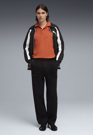 Jonge persoon staand met handen in de zakken, gekleed in een zwart-wit jack, roestoranje polo, zwarte broek en zwarte sneakers tegen een grijze achtergrond.