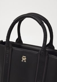 Zwarte leren handtas met dubbele korte handvatten, stoffen schouderriem, gouden logo-accent, gestructureerd ontwerp en gladde textuur.