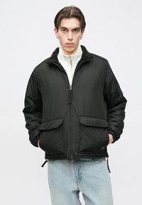 Casaco acolchoado preto com fecho de correr frontal, dois bolsos frontais e gola alta. Forro interior texturizado, punhos elásticos e detalhe de cordão na bainha.