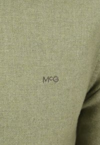 Olivengroene gebreide trui met een zachte textuur, voorzien van de geborduurde letters "McG" rechtsonder. Eenvoudig ontwerp, zonder patronen of accenten.