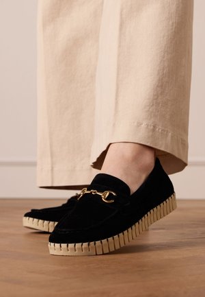 Zwarte fluwelen loafers met een gouden kettingaccent, met een gestructureerde crèmekleurige zool. Gemaakt met lichtgekleurde, loszittende broeken.
