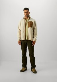 Casaco de lã creme com bolso castanho no peito, usado por cima de uma camisa verde clara, combinado com calças verde-oliva e sapatos de caminhada castanho-claro.