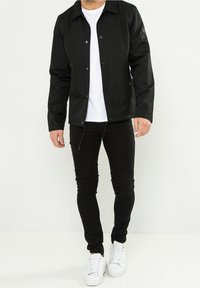 Czarna kurtka zapinana na napy z kołnierzykiem, noszona na białym t-shircie. W połączeniu z czarnymi rurkami i białymi sneakersami. Gładka tekstura materiału.