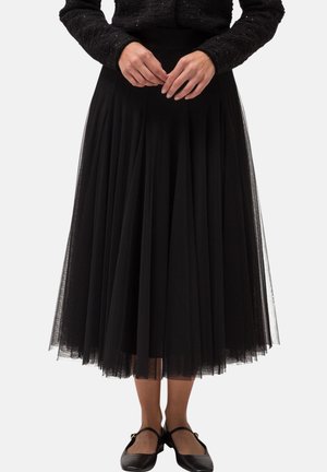 Femme portant une longue jupe en tulle noire, un pull texturé noir et des chaussures Mary Jane noires, debout les mains jointes devant elle.