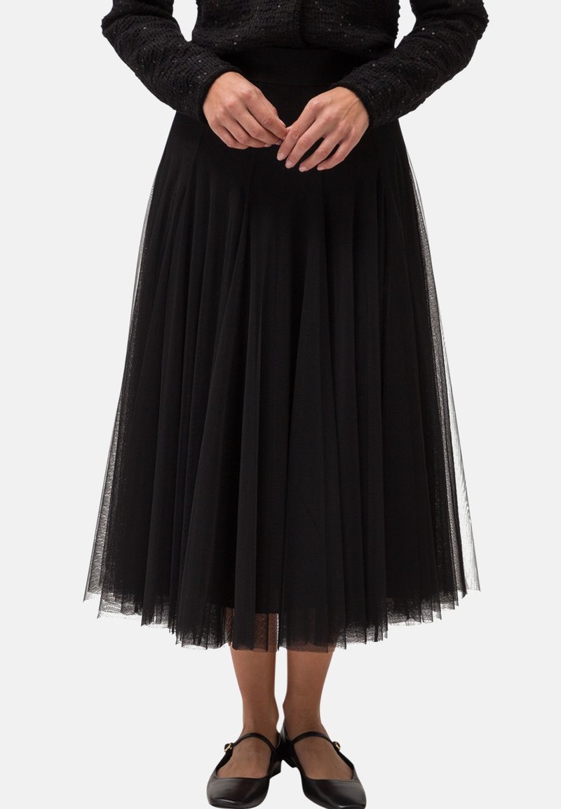 Femme portant une longue jupe en tulle noire, un pull texturé noir et des chaussures Mary Jane noires, debout les mains jointes devant elle.