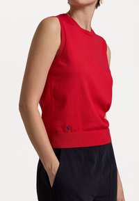 Frau in einem ärmellosen roten Stricktop mit einem kleinen marineblauen Logo nahe dem Saum, kombiniert mit schwarzen Hosen, die eine Hand in der Tasche hat.