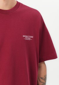 Burgunderfarvet bomulds t-shirt med runde halsudskæring, korte ærmer og hvid tekst på brystet, der står "ORIGINALS STUDIO Athletic Association."