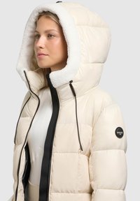 Gewatteerd donzen jack in roomkleur met een capuchon gevoerd met nepbont, zwarte ritssluiting en een logopatch op de linker mouw. Gladde textuur, boxy model.