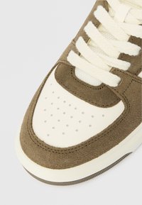 Detalj na vrhu tenisice u smeđem nubuku i bijeloj koži s kremastim vezicama i perforacijama na bijeloj potplati.