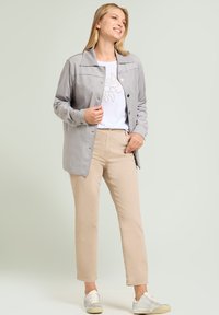 Chaqueta de botones gris claro con una textura suave, llevada sobre una camiseta blanca con un diseño beige simple, combinada con pantalones beige y zapatillas.