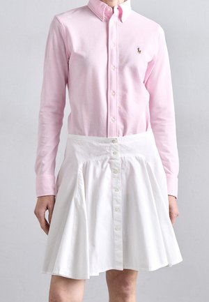 Person wearing a light pink buttoned shirt with long sleeves and a white high-waisted buttoned skirt against a plain background.