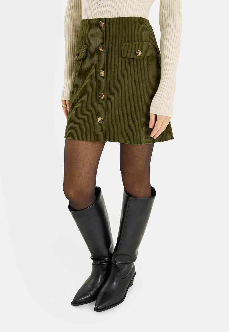 Mini-jupe en velours côtelé vert avec boutons devant et deux poches à rabat, associée à des bottes en cuir noires montant jusqu'aux genoux.