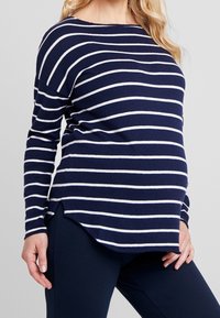 Femme enceinte portant un haut à manches longues rayé bleu marine et blanc avec des leggings de maternité bleu marine, se tenant devant un fond uni.