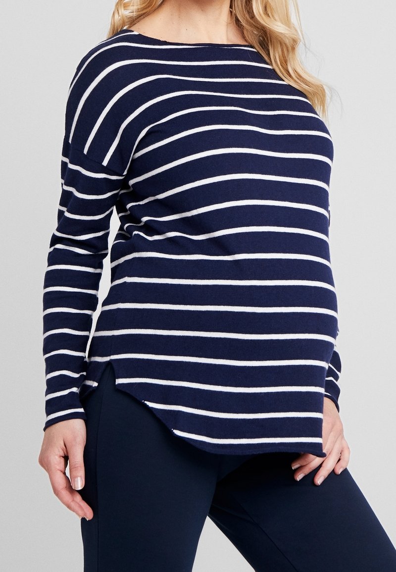 Femme enceinte portant un haut à manches longues rayé bleu marine et blanc avec des leggings de maternité bleu marine, se tenant devant un fond uni.