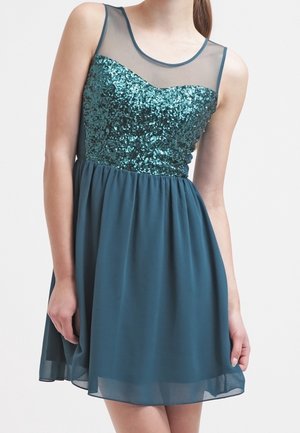 Robe bleu sarcelle sans manches avec corsage à paillettes, encolure transparente et jupe fluide en chiffon portée par une personne debout.