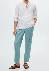 Weißes Leinenhemd mit langen Ärmeln und Stehkragen, kombiniert mit hellblauen, strukturierten, locker sitzenden Hosen und braunen Slip-on-Sandalen.