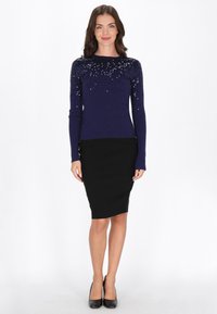 Tengerés színű, flitteres pulóver hosszú ujjakkal, fekete, testhez simuló ceruzaszoknya és fekete, hegyes orrú magassarkú cipő. Simább anyag, csillogó felső dizájn és testhez simuló stílus.
